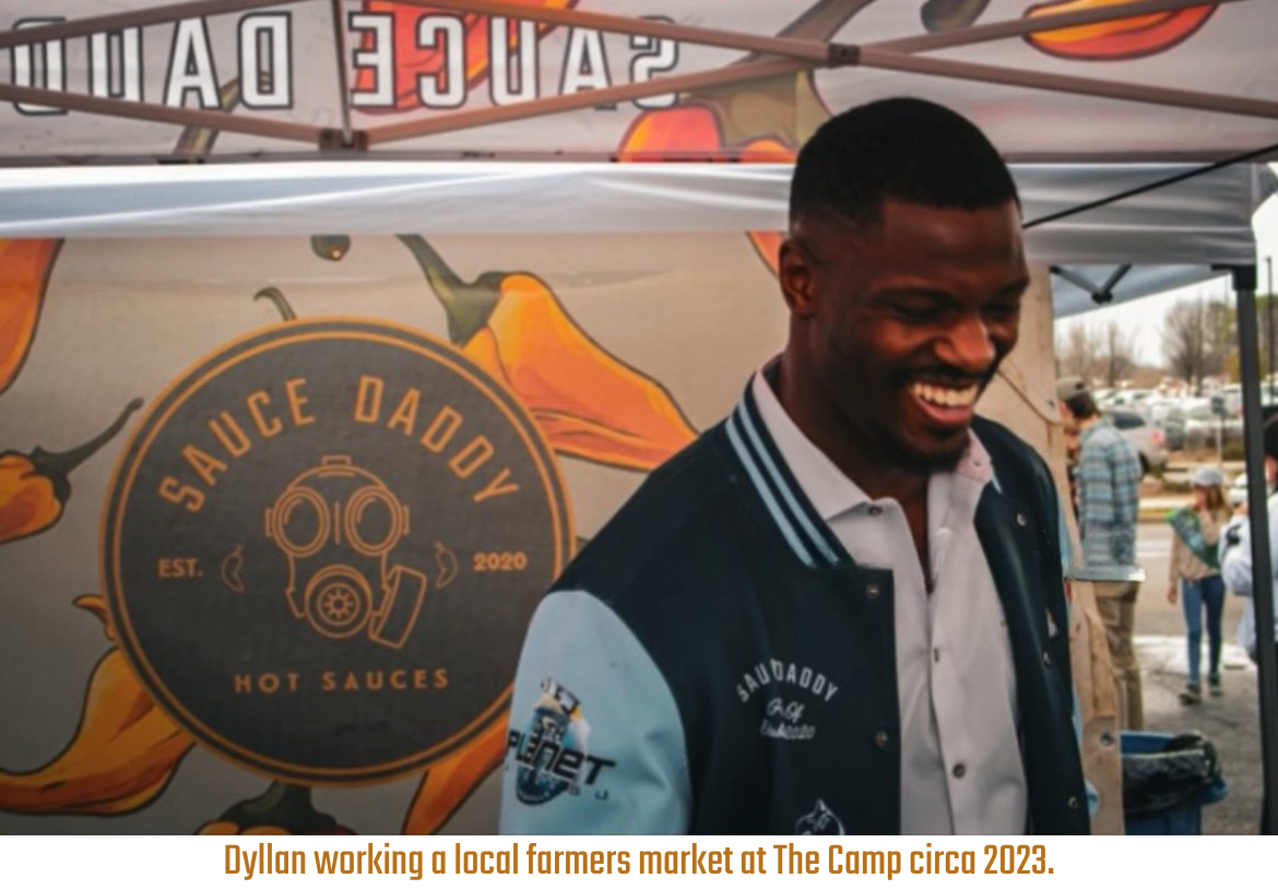 Company Founder Dyllan standing in front of a 'Sauce Daddy' banner in hot sauce stand at a farmers market.