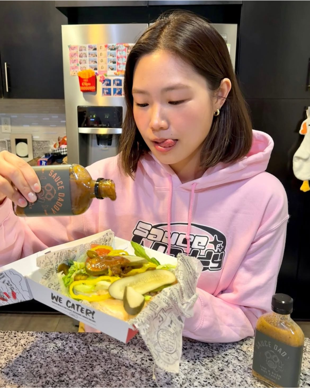 A Sauce Daddy influencer wearing a pink Sauce Daddy hoodie pouring Sauce Daddy hot sauce over a burger in a kitchen.